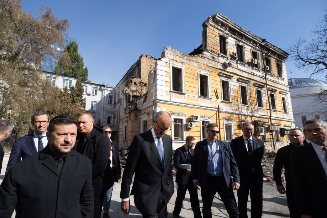 Volodymyr Zelenskyy and Friedrich Merz walk past a building in Kyiv damaged by a Russian strike, surrounded by officials.