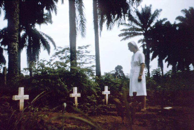A nun in a white habit walks among simple white crosses marking graves beneath palm trees.
