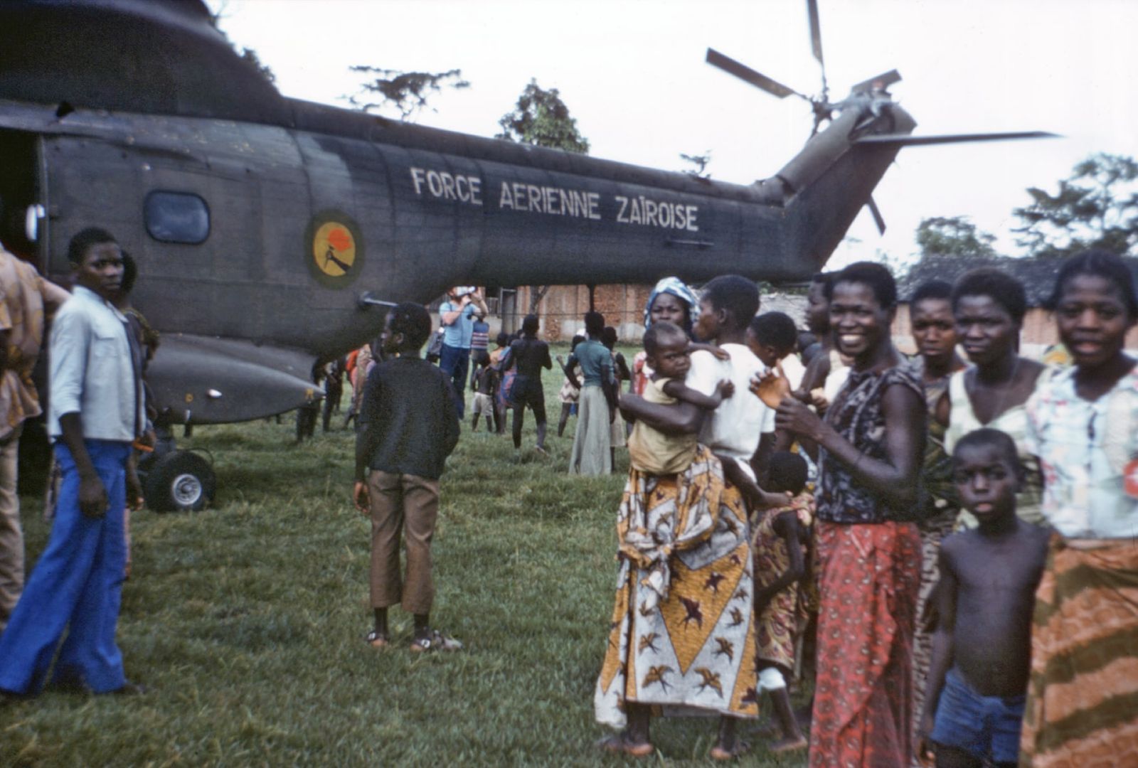 A large military helicopter marked Force Aérienne Zaïroise on a grass field, surrounded by local residents.
