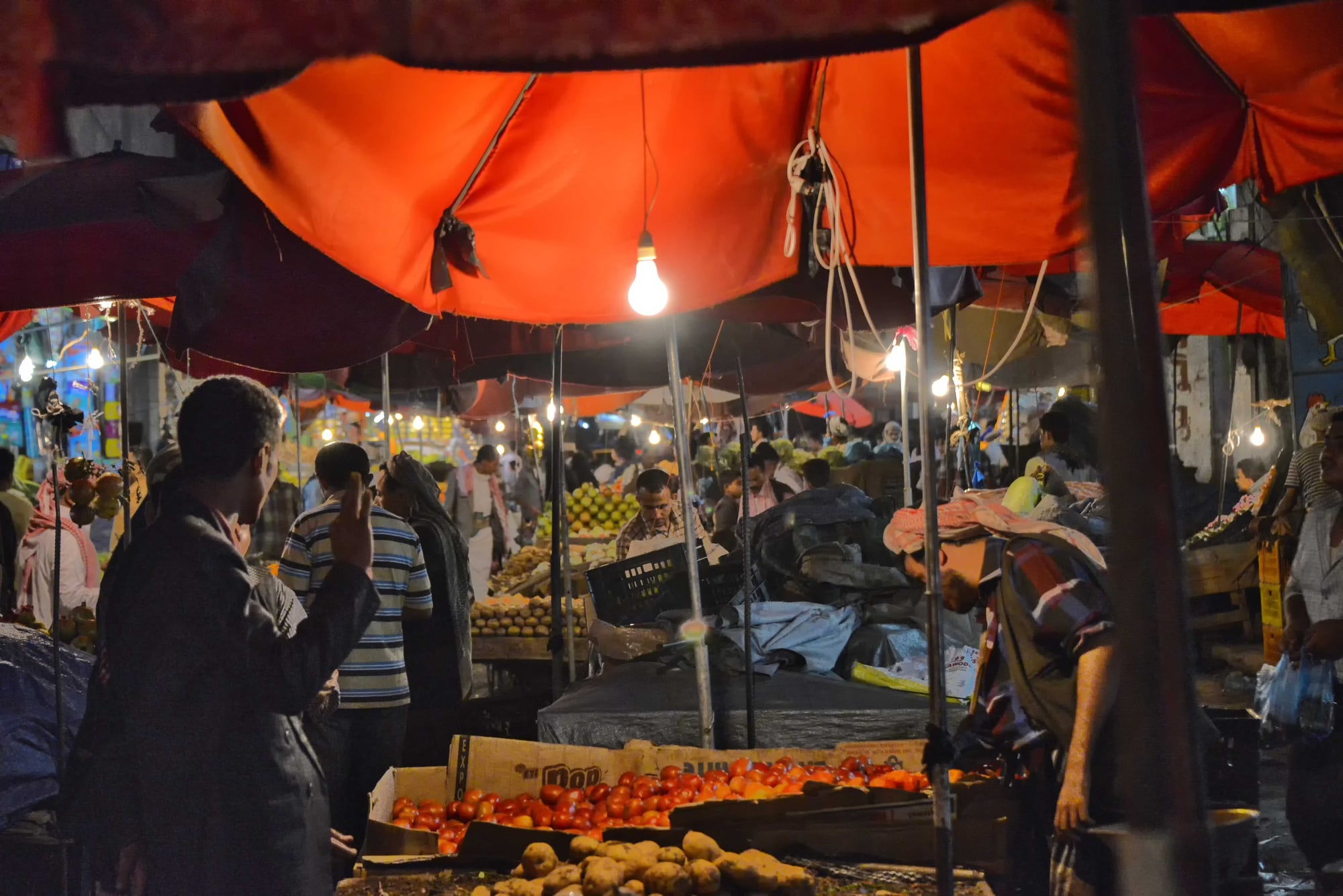 A busy evening market in Hodeidah, with fruit stalls under orange awnings lit by bare bulbs.