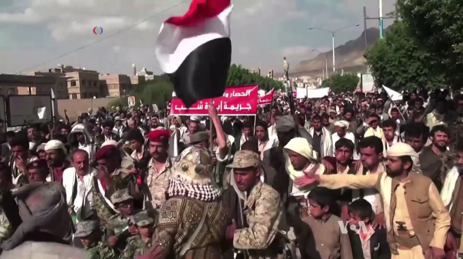 A large crowd of Houthi supporters, some armed, march through a Sanaa street holding a Yemeni flag and banners.