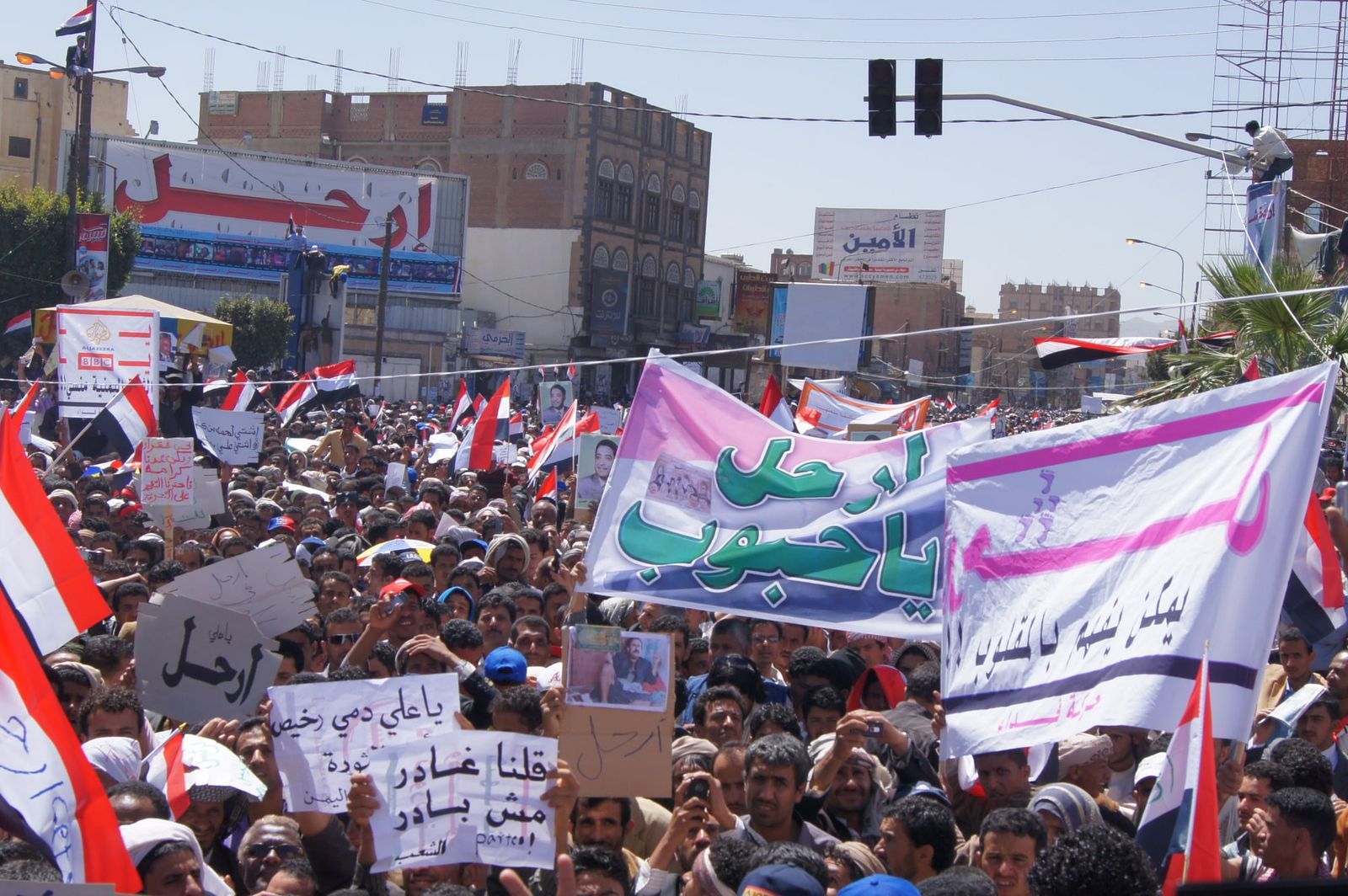 A dense crowd of protesters with Yemeni flags and banners in Arabic fills a Sanaa street.