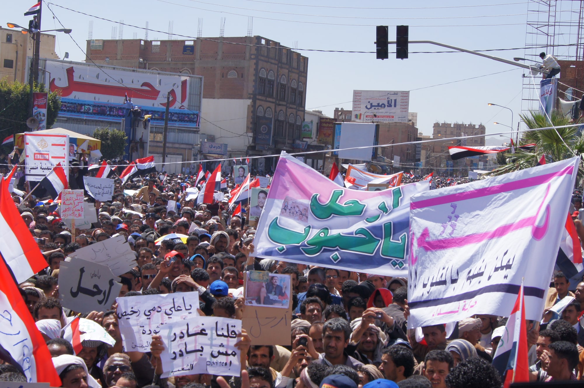 A dense crowd of protesters with Yemeni flags and banners in Arabic fills a Sanaa street.