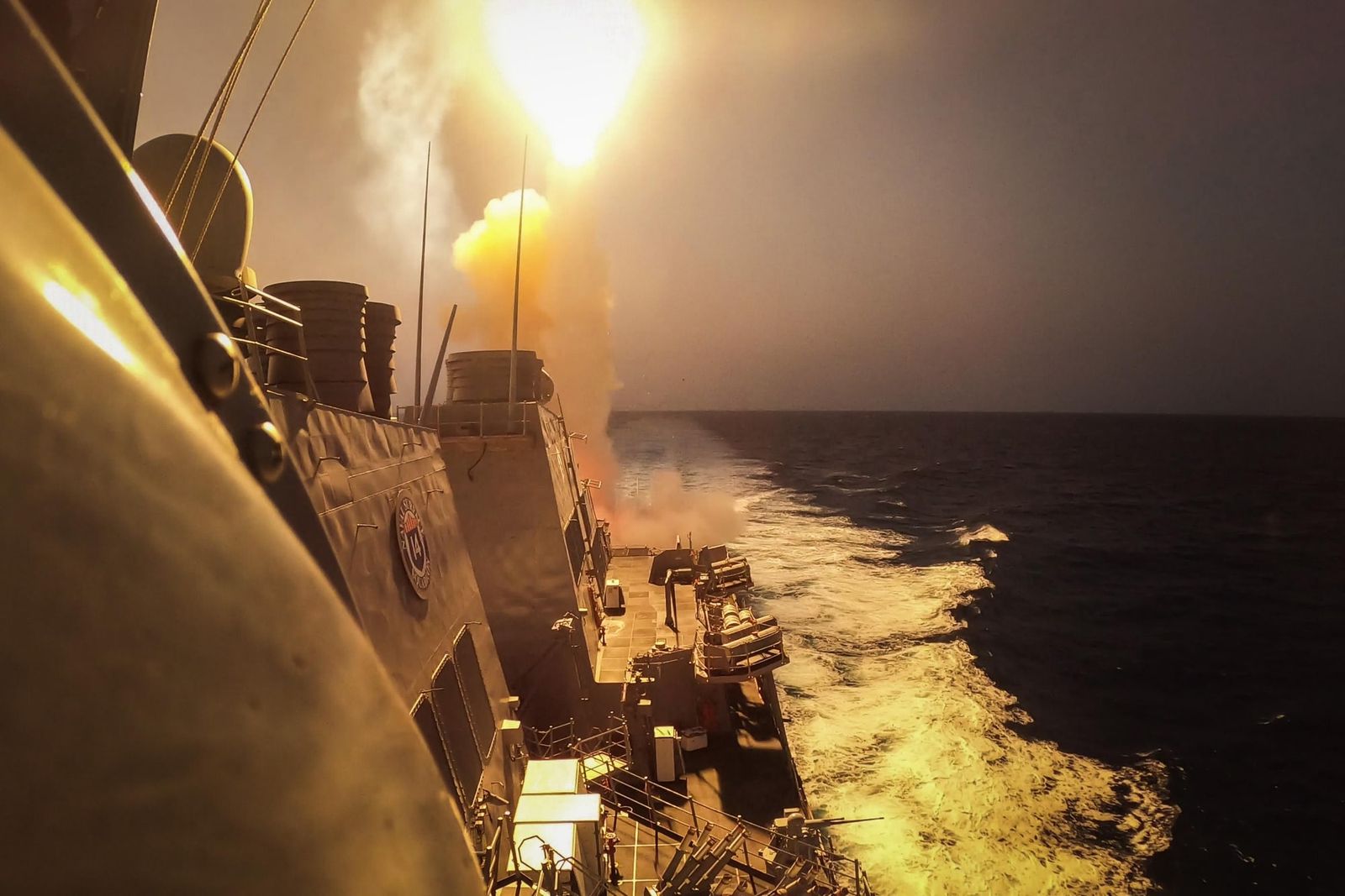 A US Navy destroyer fires a missile at sea, its launch lighting up the sky.
