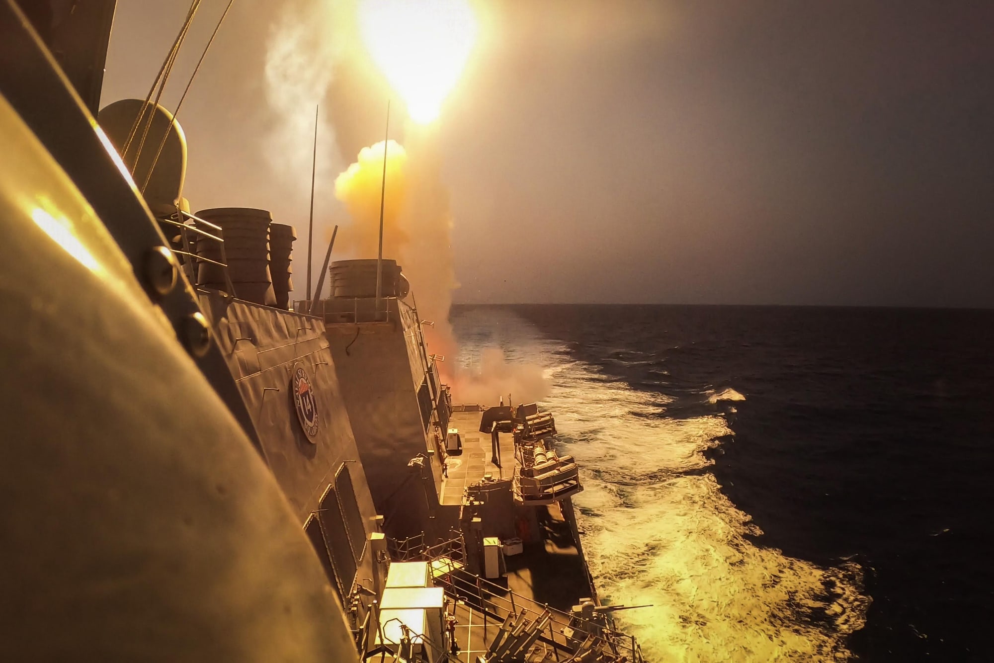 A US Navy destroyer fires a missile at sea, its launch lighting up the sky.