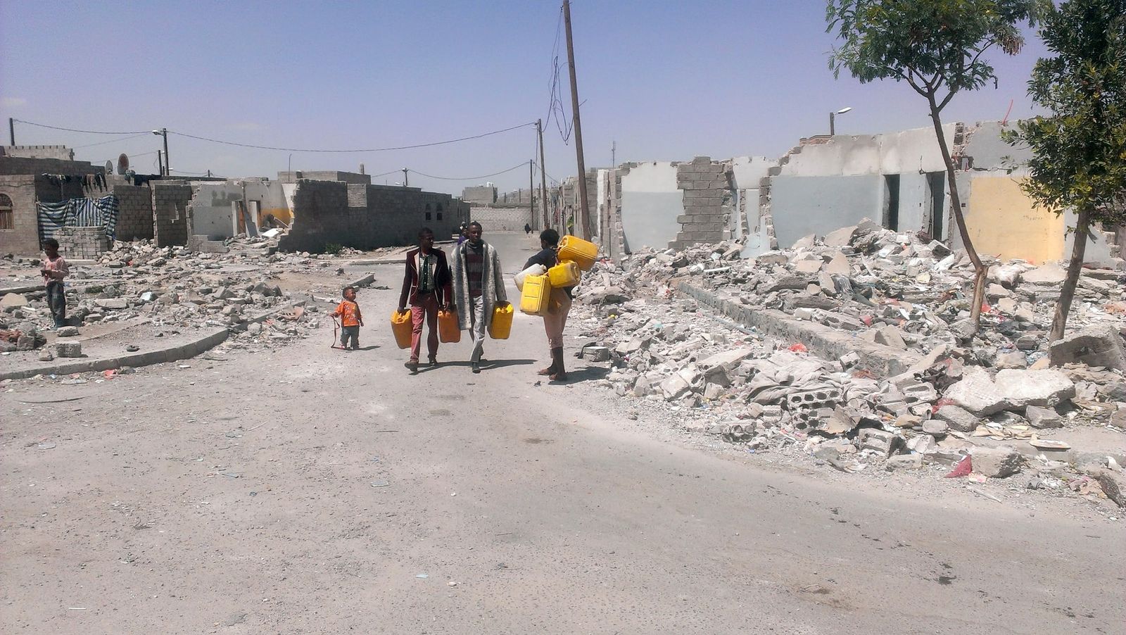 Men and a child carry yellow water containers along a street lined with damaged buildings.