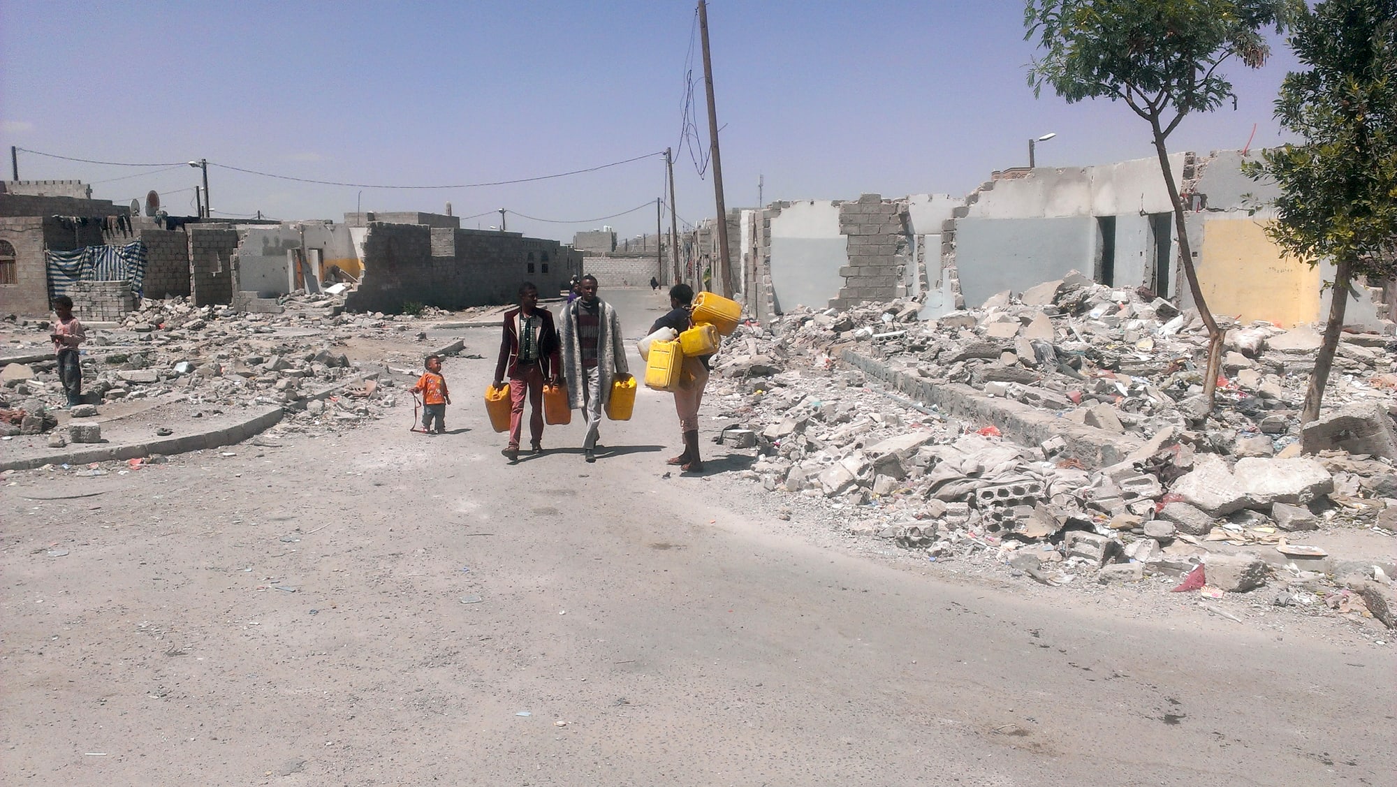 Men and a child carry yellow water containers along a street lined with damaged buildings.