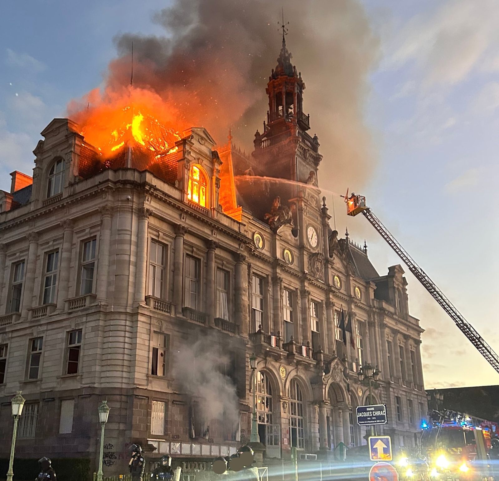 Limoges city hall on fire on the evening of 17 October 2026, flames through the roof beside the clock tower, with a fire engine's ladder spraying water.