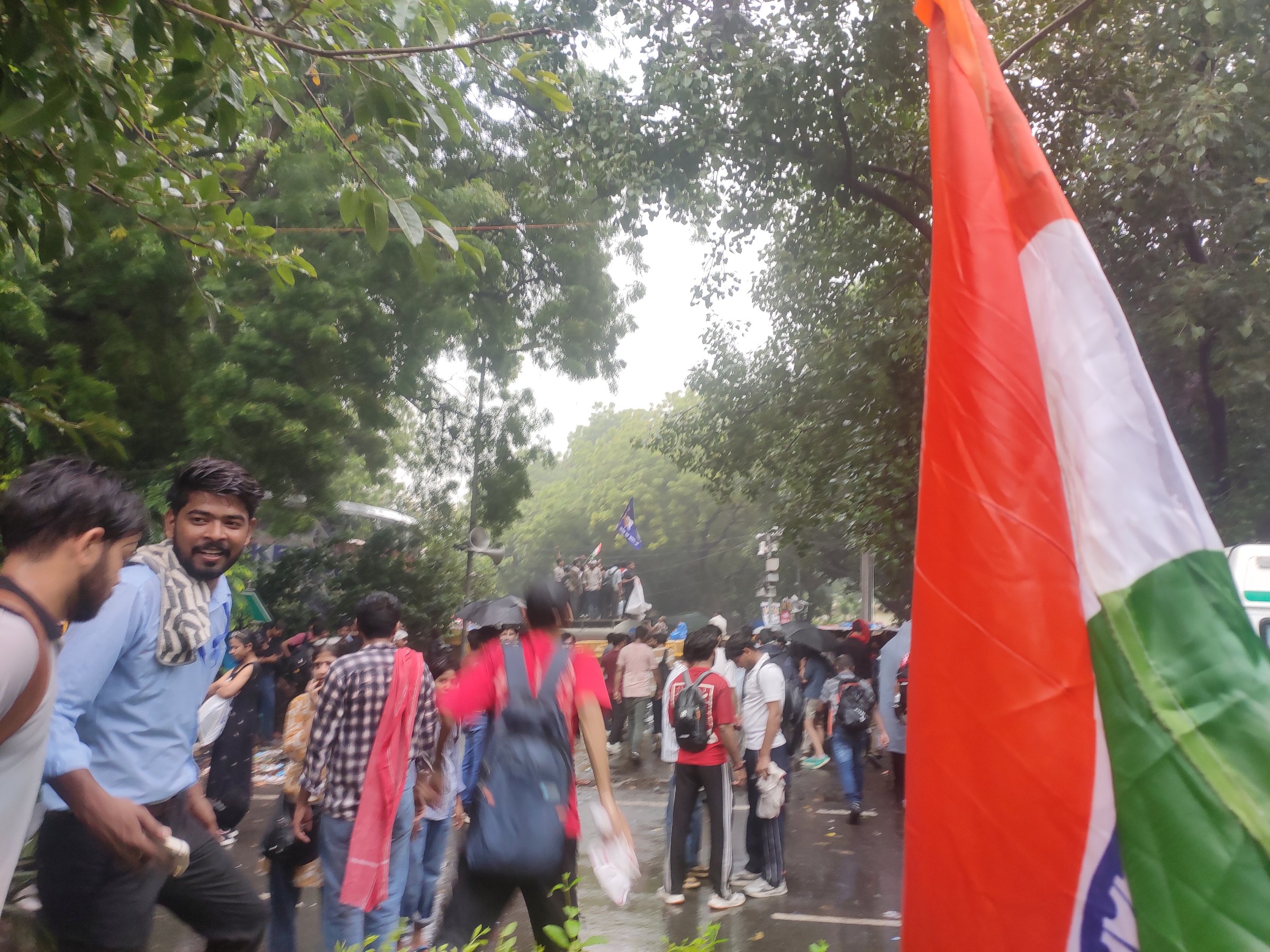 Young protesters walk along a wet, tree-lined street in New Delhi, an Indian flag in the foreground and more flags raised on a vehicle ahead