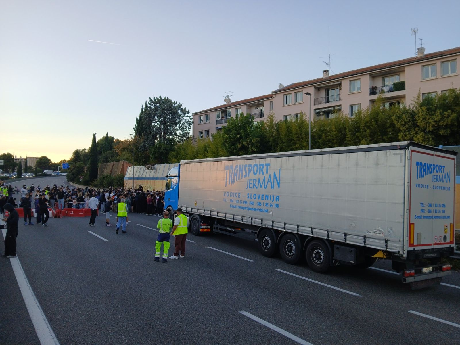 Protesters stand across the lanes of a motorway at dusk beside a stopped lorry, with workers in high-visibility vests