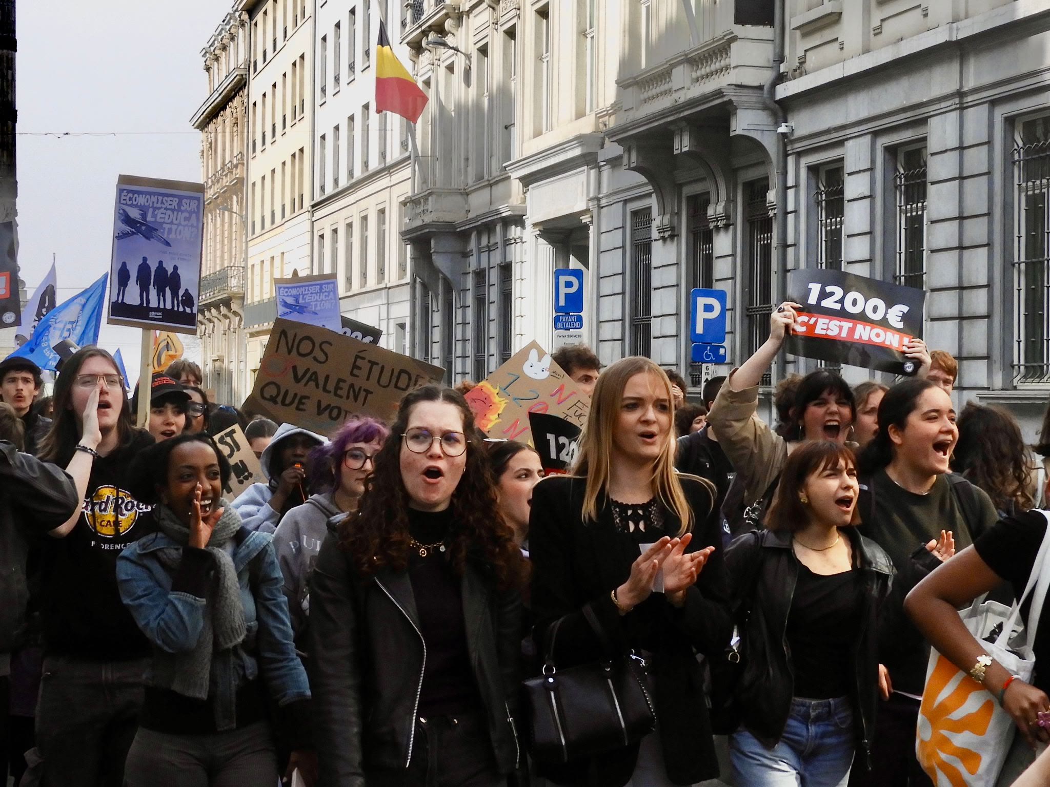 Young women march through a Brussels street shouting, one holding a placard reading "1200€ c'est non"