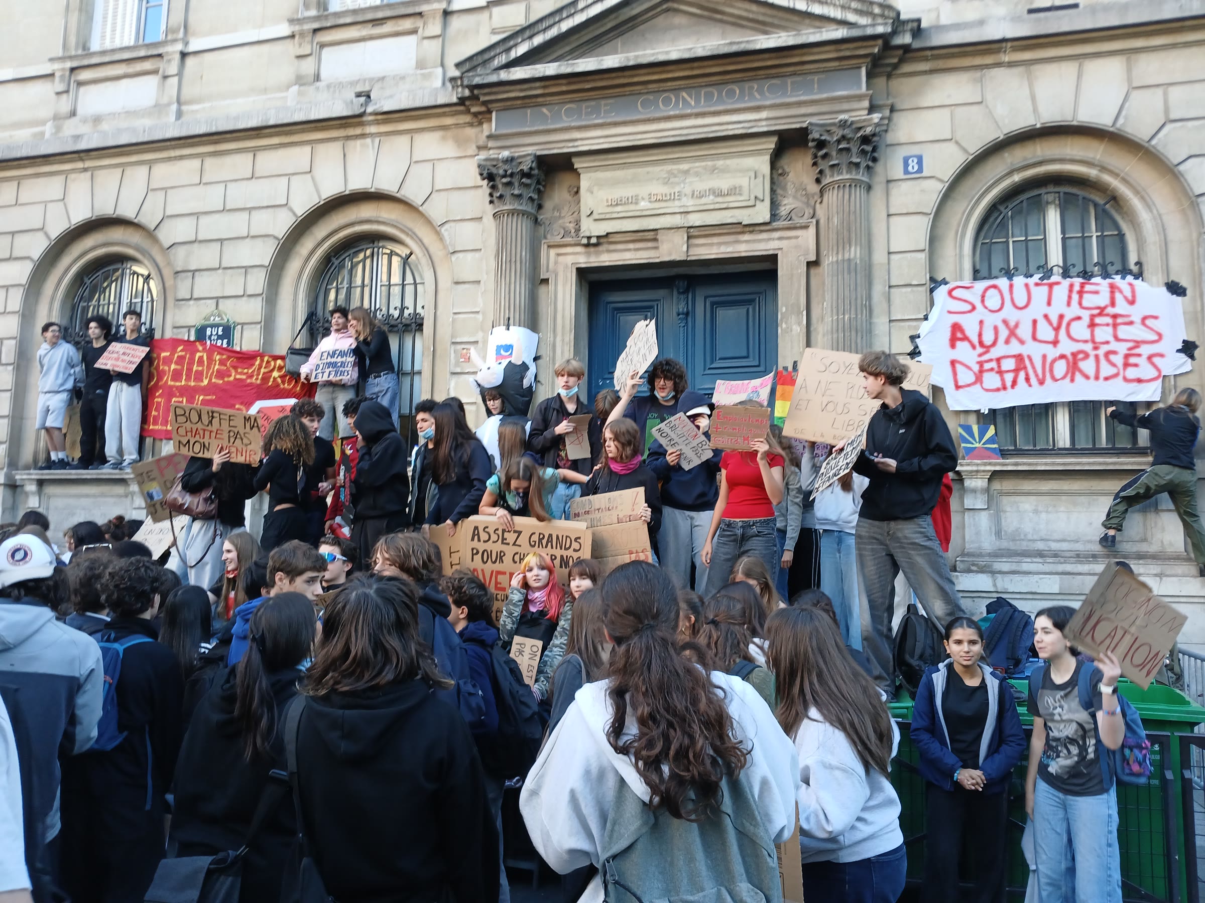 Teenage students crowd the steps of Lycée Condorcet in Paris holding cardboard placards, under a banner reading "Soutien aux lycées défavorisés"