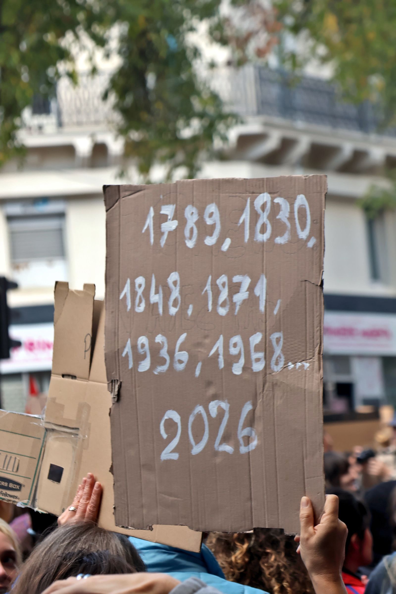 A cardboard placard held up in a Paris crowd lists the years 1789, 1830, 1848, 1871, 1936, 1968 and 2026