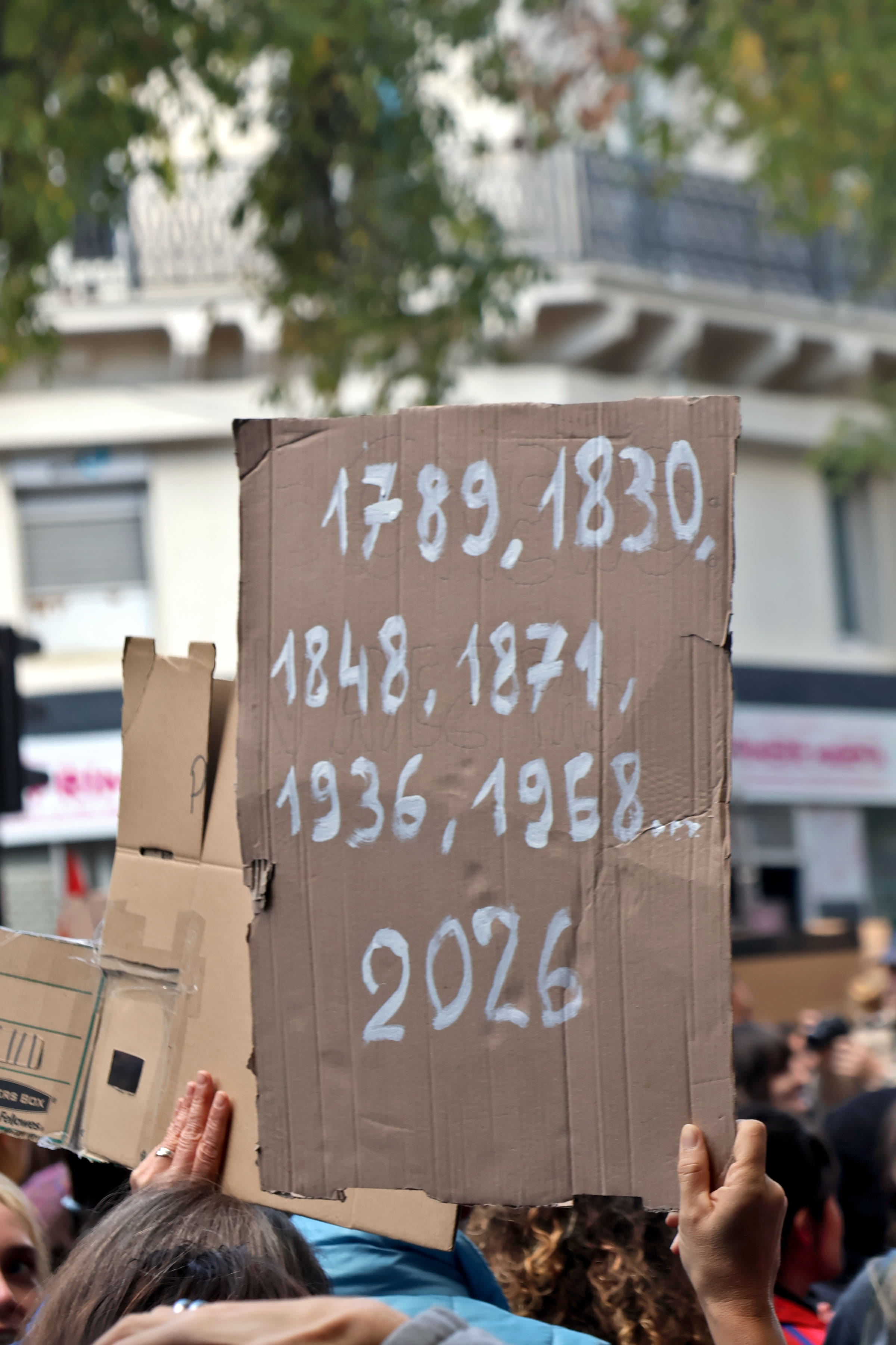 A cardboard placard held up in a Paris crowd lists the years 1789, 1830, 1848, 1871, 1936, 1968 and 2026