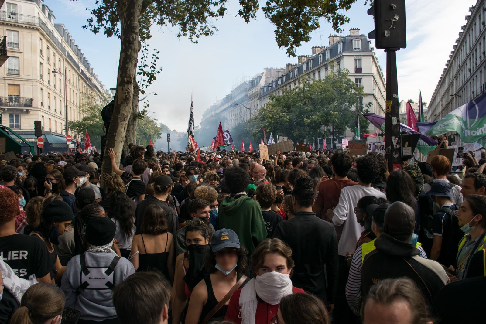 A dense crowd of young marchers fills a tree-lined Paris boulevard, flags and smoke in the distance