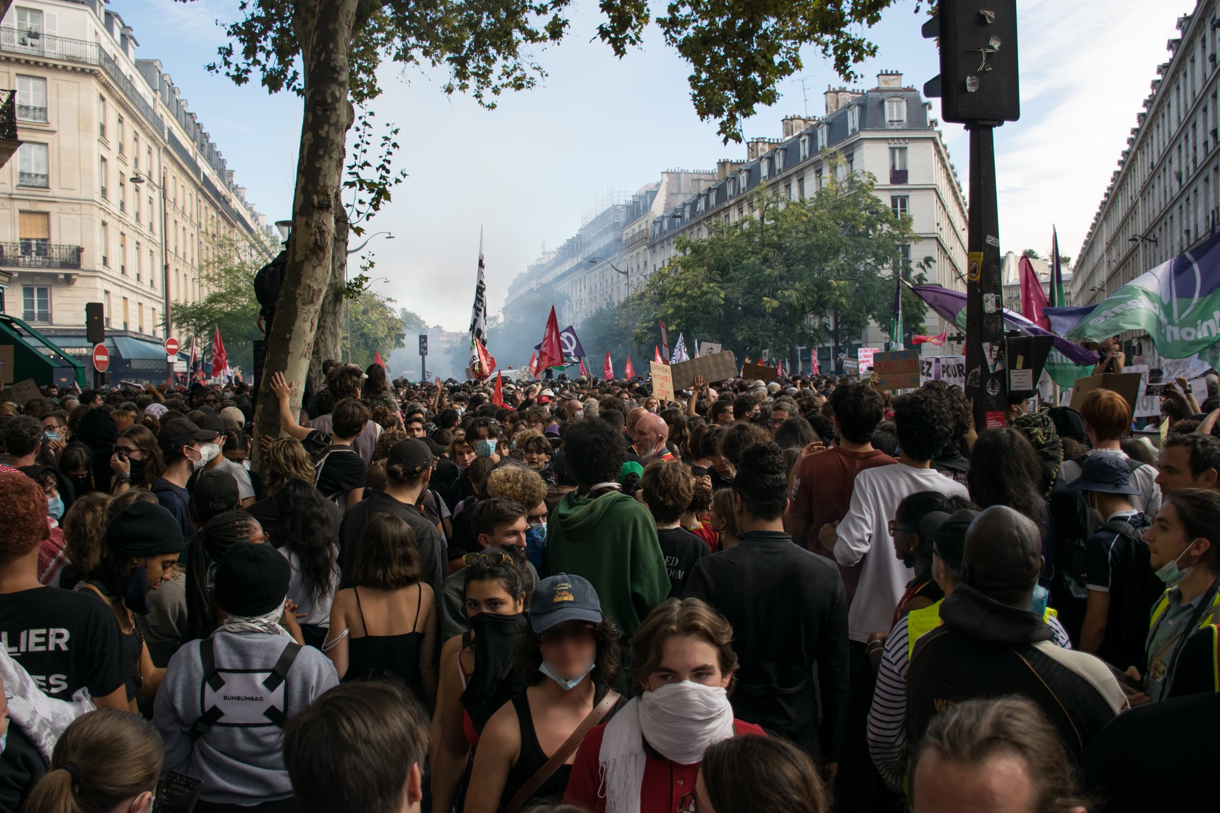 A dense crowd of young marchers fills a tree-lined Paris boulevard, flags and smoke in the distance