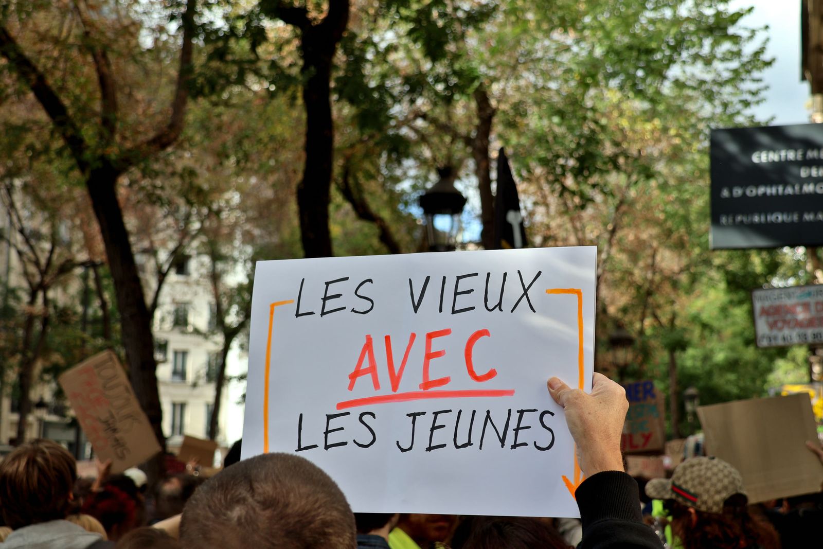 A white placard reading "Les vieux avec les jeunes" held above a crowd under trees