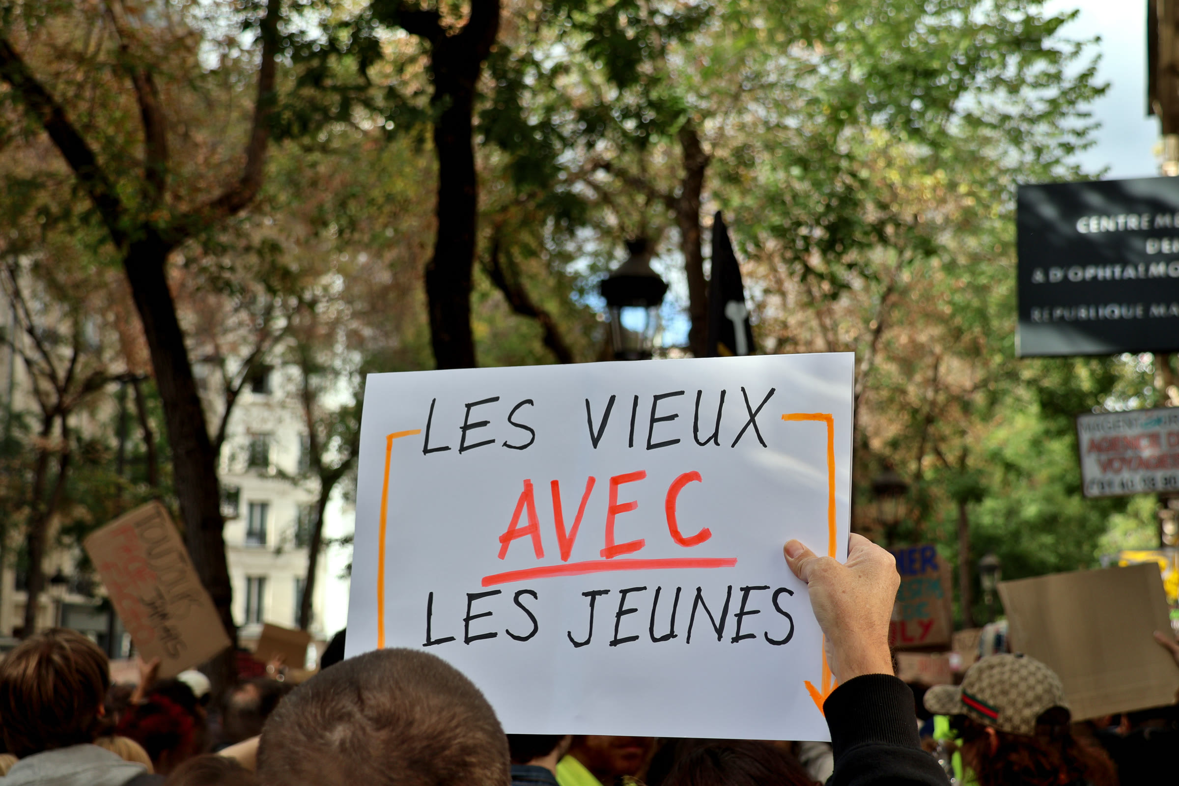 A white placard reading "Les vieux avec les jeunes" held above a crowd under trees