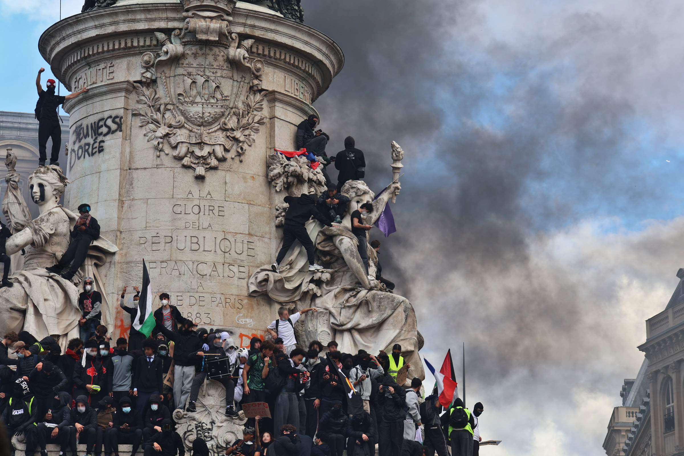 Protesters dressed in black climb the monument in the Place de la République in Paris as dark smoke rises behind it
