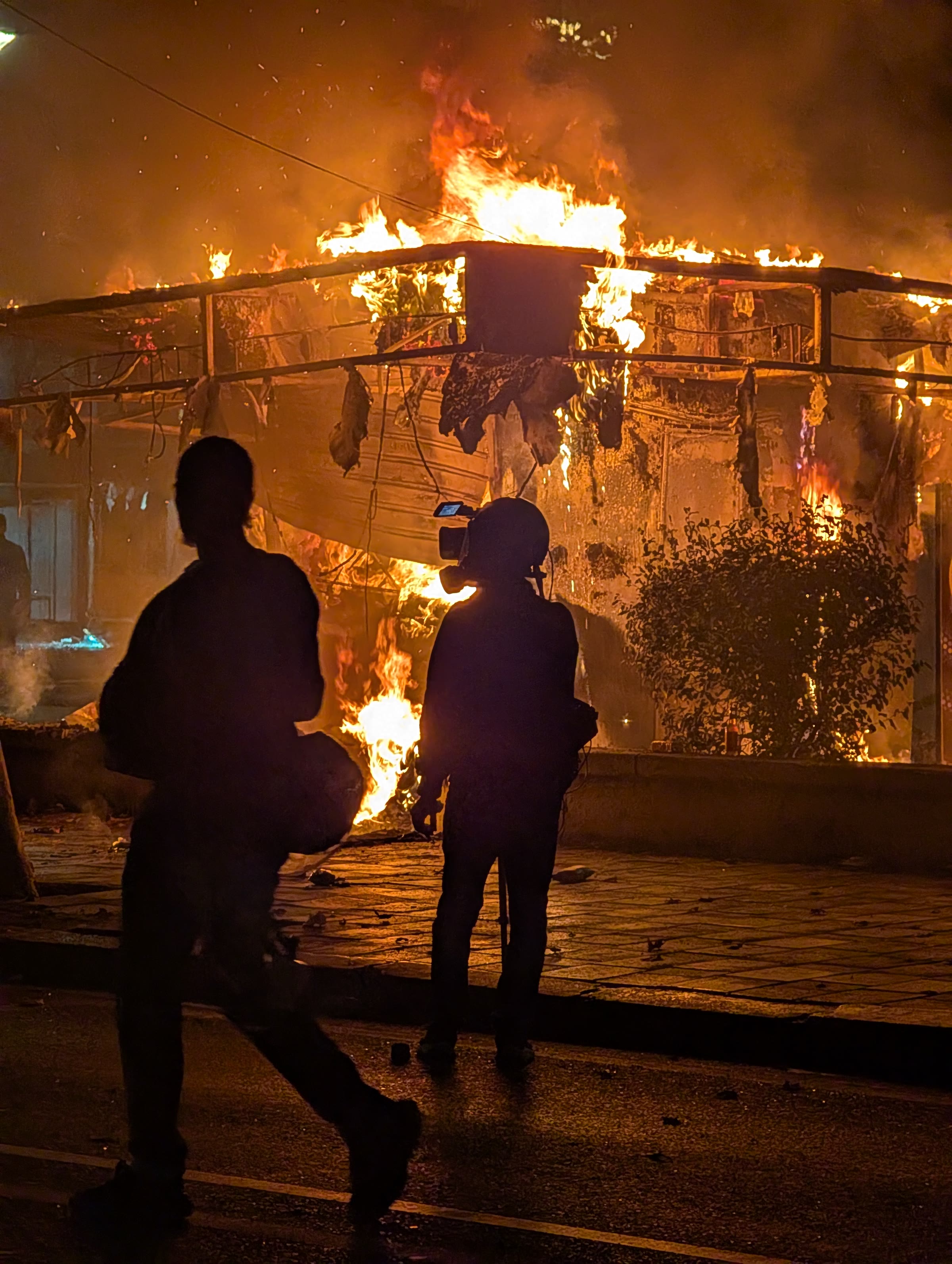 Silhouetted figures stand before a fire burning against railings at night in Tirana