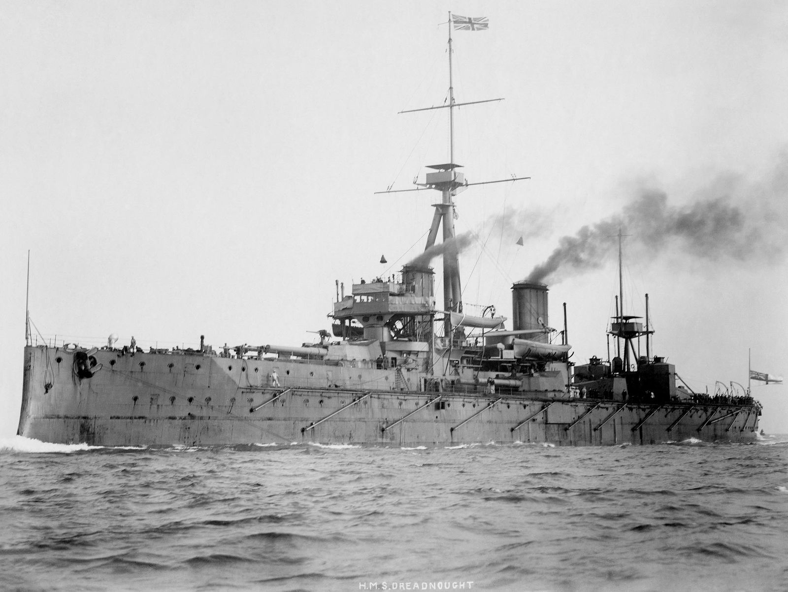 HMS Dreadnought at sea in 1906, a long grey battleship with two tall masts and smoke rising from its funnels.