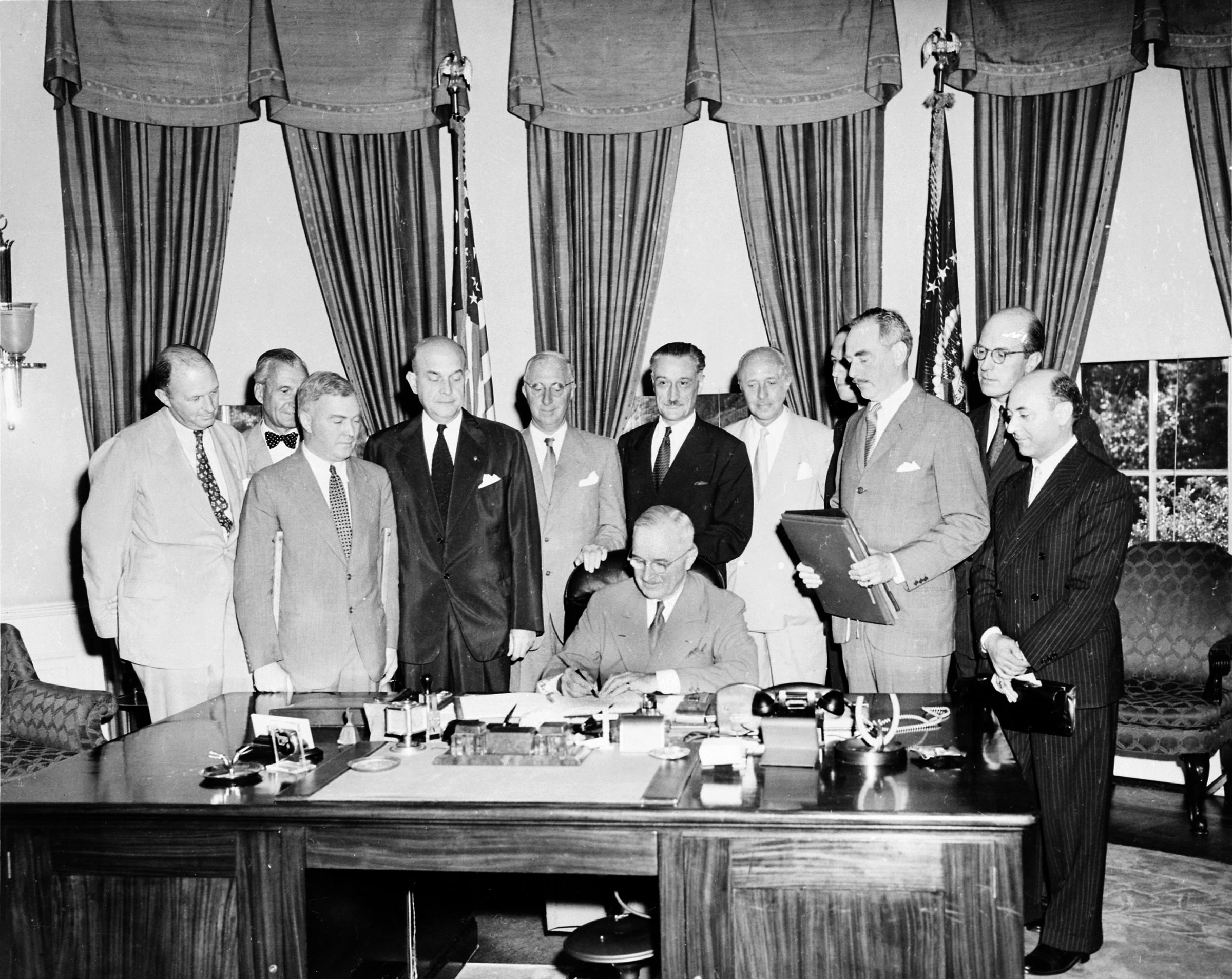President Harry Truman signs a document at a desk, surrounded by officials, as the North Atlantic Treaty comes into force in August 1949.