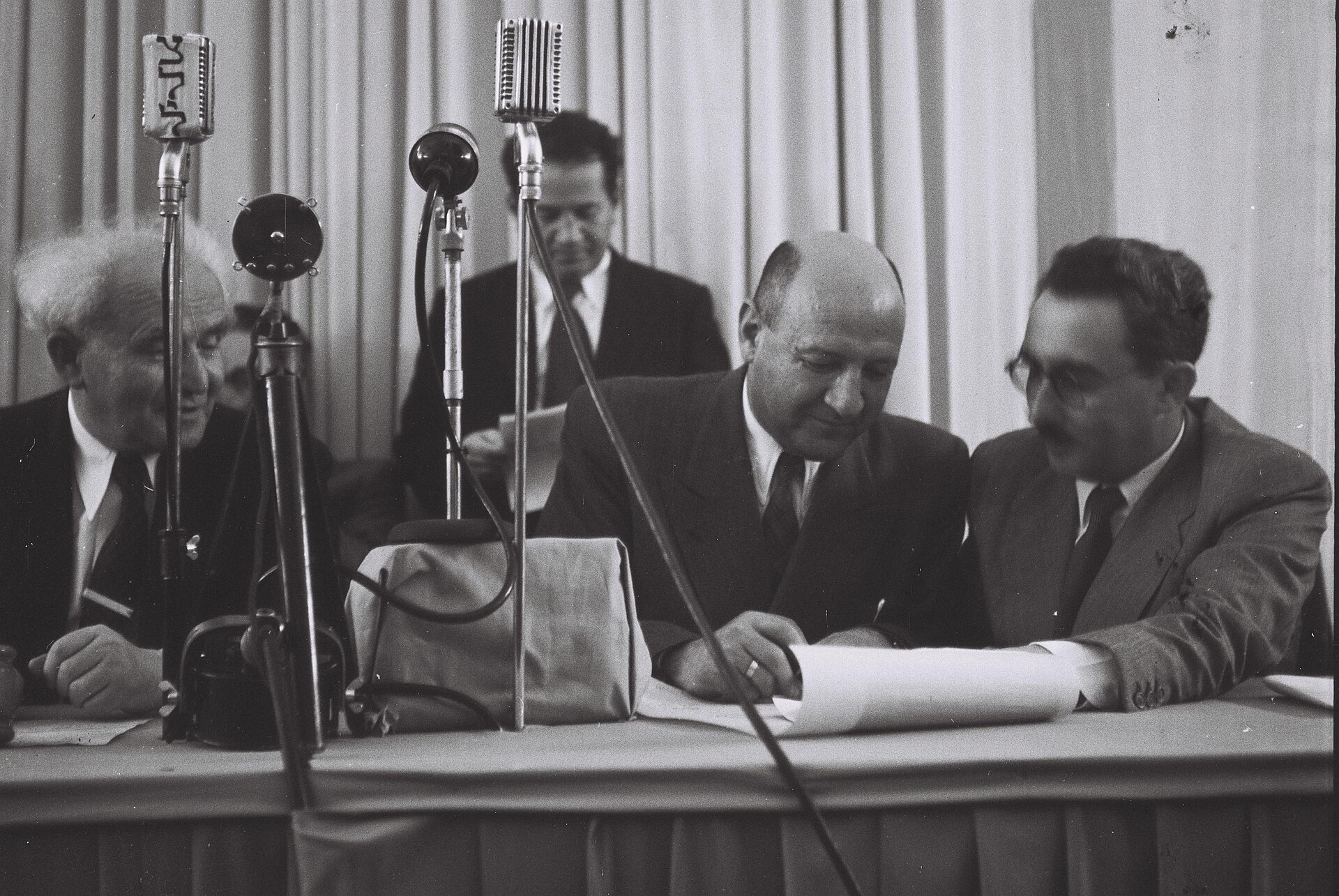 Men in suits seated at a table with microphones, one signing a document as David Ben-Gurion looks on