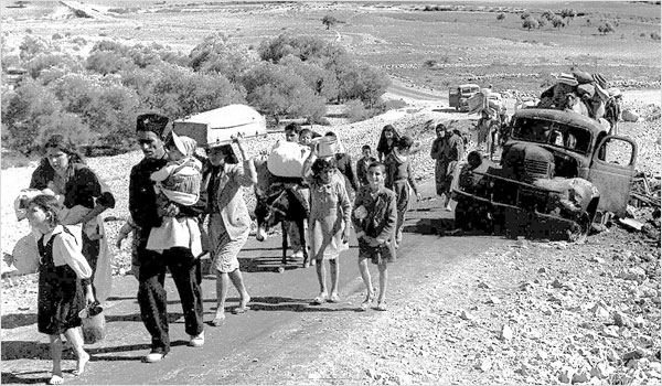 Women, men and children walking along a rough road carrying bundles, past a broken-down truck