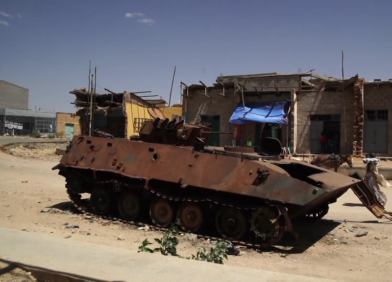 A burnt-out armoured vehicle on a dusty roadside in front of damaged shops
