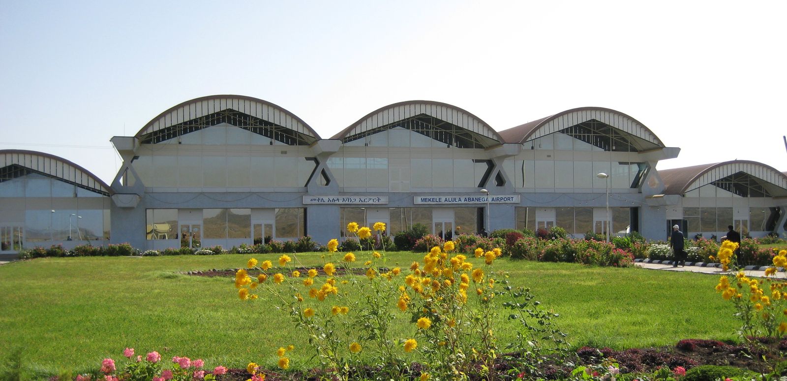 A low terminal building with curved roofs behind a lawn and flowerbeds