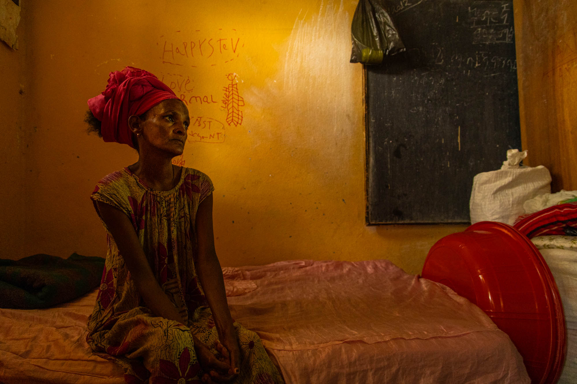 A woman in a red headscarf sits on a bed in a classroom, a blackboard behind her