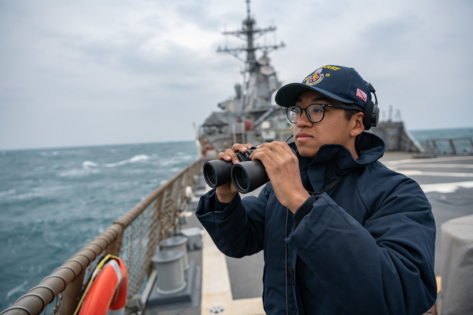 A US Navy sailor with binoculars on lookout on the flight deck of a destroyer at sea