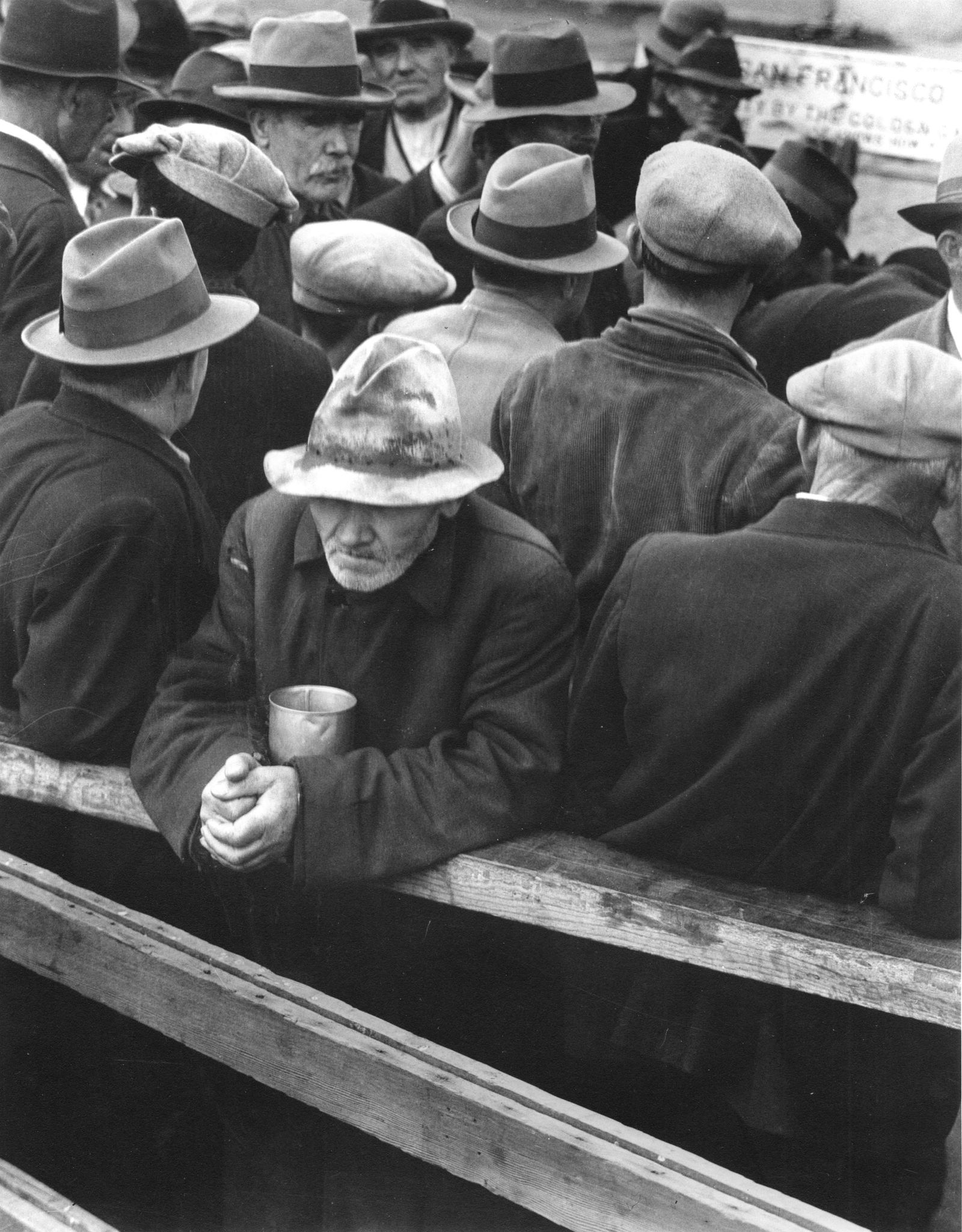 A crowd of men in hats and coats queue at a breadline in San Francisco in 1932; one man in the foreground leans on a rail holding a tin cup.