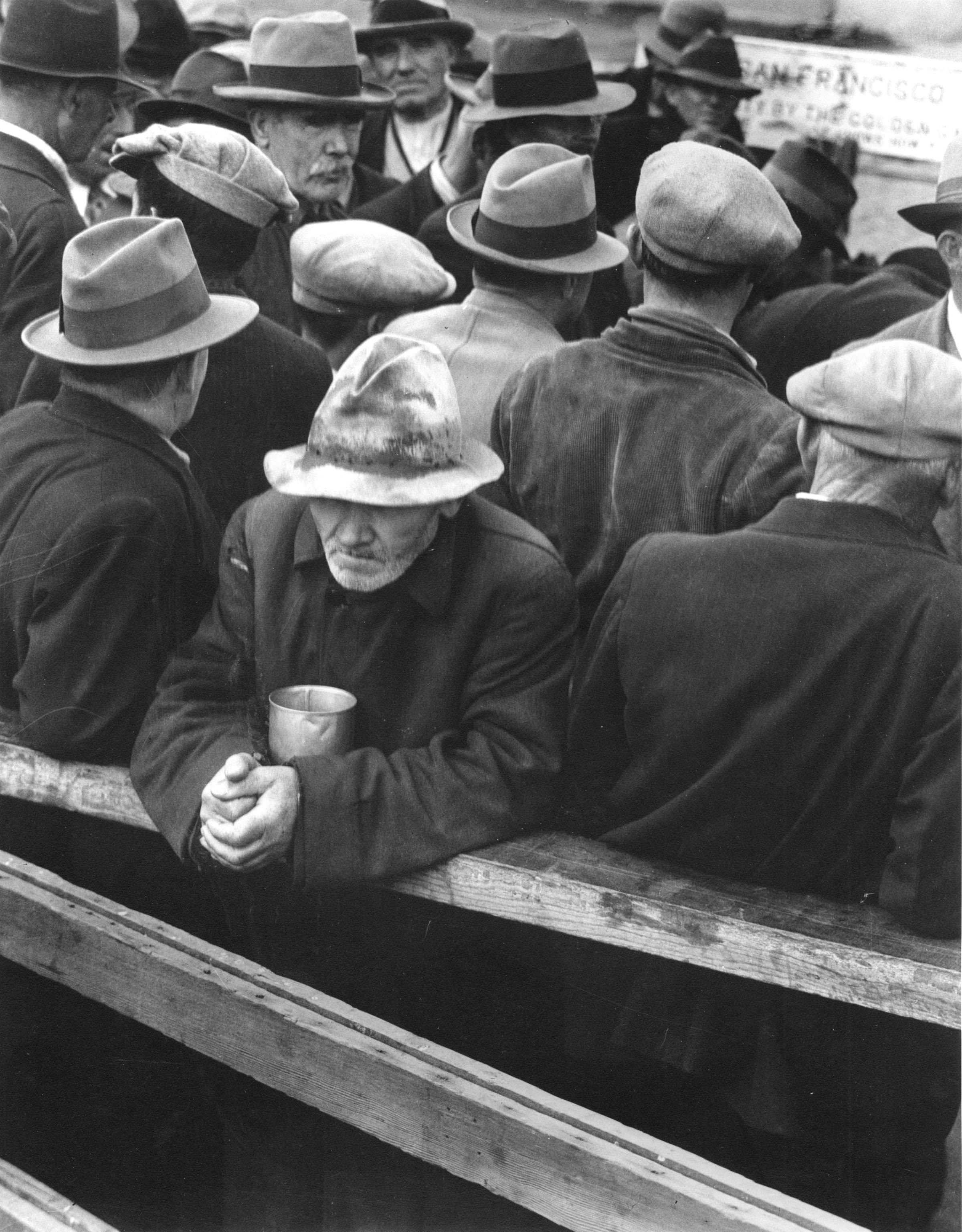 A crowd of men in hats and coats queue at a breadline in San Francisco in 1932; one man in the foreground leans on a rail holding a tin cup.